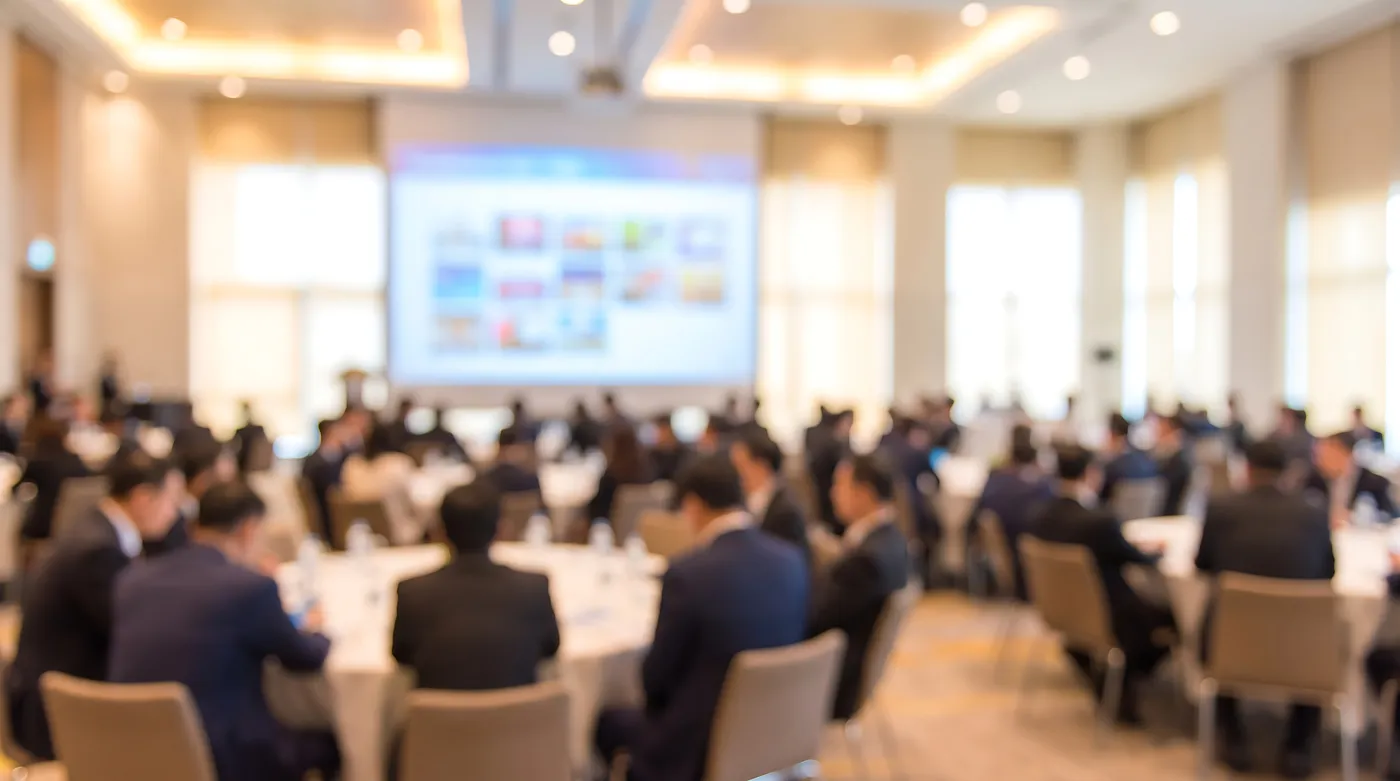 Soft-focus view of conference attendees seated at tables facing a presentation screen.
